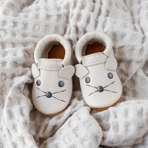Peut inclure: Une paire de chaussons en cuir gris clair pour b&eacute;b&eacute; avec des oreilles de souris et des yeux et des moustaches brod&eacute;s noirs.