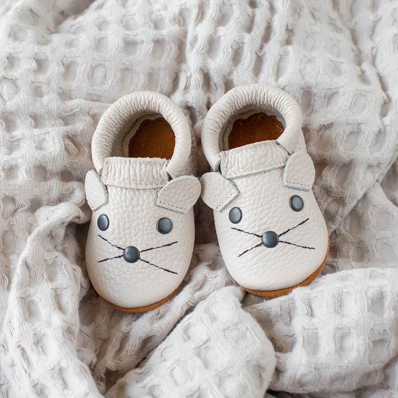 Peut inclure: Une paire de chaussons en cuir gris clair pour b&eacute;b&eacute; avec des oreilles de souris et des yeux et des moustaches brod&eacute;s noirs.
