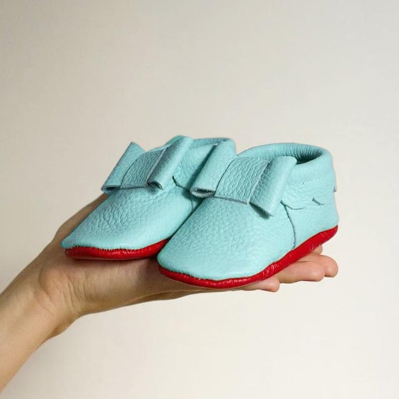 Pu&ograve; includere: Un paio di mocassini per bambini di colore azzurro chiaro con un bordo rosso. I mocassini hanno un dettaglio a fiocco sulla parte superiore.