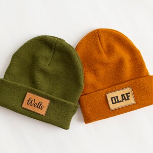 May include: Two knitted beanies with leather patches. One beanie is olive green with the word "Wells" on the patch. The other beanie is orange with the word "OLAF" on the patch.