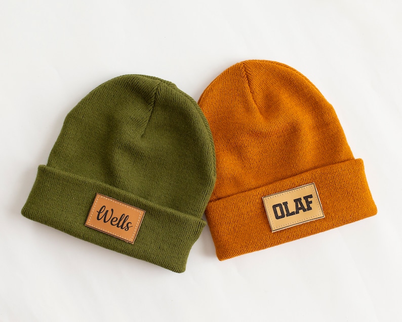 May include: Two knitted beanies with leather patches. One beanie is olive green with the word "Wells" on the patch. The other beanie is orange with the word "OLAF" on the patch.