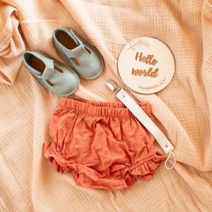 Puede incluir: Un conjunto de ropa de beb&eacute; que incluye un par de zapatos de beb&eacute; azul claro, un par de bombachos de color naranja con volantes y un letrero de madera que dice "Hello world".