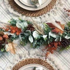 May include: A decorative table centerpiece featuring a mix of artificial greenery, brown pinecones, orange autumn leaves, and white pampas grass. The arrangement is placed on a light-colored striped tablecloth between two round woven placemats.