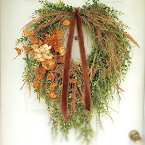 May include: A fall-themed wreath with a mix of greenery, orange leaves, and faux pumpkins. A cluster of flowers and pinecones adds texture. A brown ribbon hangs down, creating a rustic and seasonal decoration for a front door.