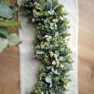May include: A half-circle artificial wreath featuring a mix of green foliage and small white flowers. The wreath includes various shades of green leaves and soft, silvery-green accents. The wreath is placed on a white, textured surface.