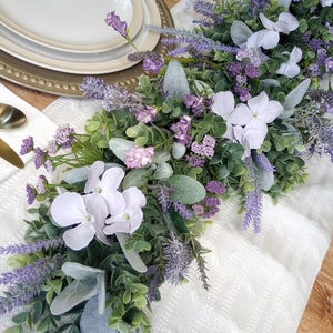 May include: A floral table runner featuring a mix of lavender, white, and green foliage. The runner is adorned with lavender sprigs, white flowers, and various green leaves, creating a natural and elegant centerpiece. The runner is placed on a white textured tablecloth.