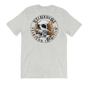 May include: Pale grey t-shirt with a circular design featuring a skull on a skateboard, with flames. The text "SLINKBONE" is above the skull, and "FEARLESS IN MOTION" below. The design is in black, white, and orange.