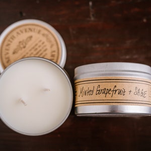 Puede incluir: Una vela en una lata de plata con una etiqueta marrón que dice "Minted Grapefruit + Sage". La vela está sobre una superficie de madera.