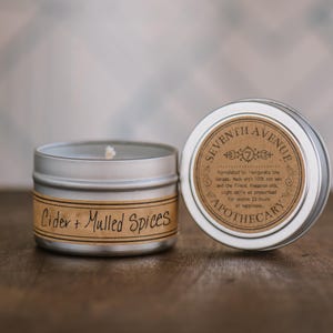 May include: A silver tin candle with a brown label that reads "Cider + Mulled Spices". The lid of the tin is silver with a brown label that reads "Seventh Avenue Apothecary". The label has a small illustration of a number seven.