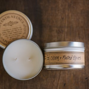 May include: Two silver candle tins with brown labels. The label on the left tin reads "Seventh Avenue" and the label on the right tin reads "Cider + Mulled Spices".