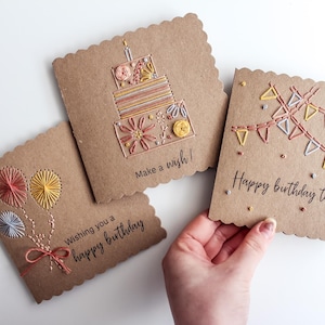 May include: Three handmade birthday cards with scalloped edges. Each card features embroidered designs: balloons, a cake, and a banner. The cards are brown with colorful thread and the text "Wishing you a happy birthday", "Make a wish!", and "Happy birthday to you".