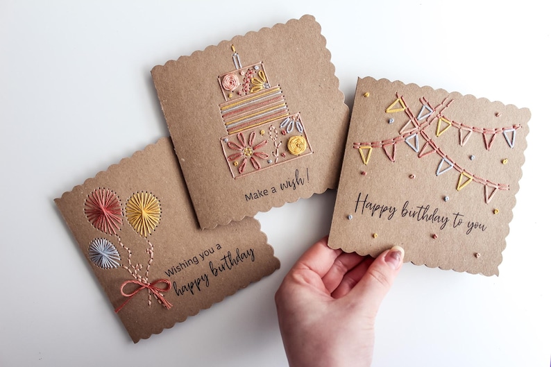 May include: Three handmade birthday cards with scalloped edges. Each card features embroidered designs: balloons, a cake, and a banner. The cards are brown with colorful thread and the text "Wishing you a happy birthday", "Make a wish!", and "Happy birthday to you".