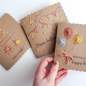 May include: Three handmade birthday cards with scalloped edges. The cards are made of brown cardstock and feature colorful embroidered designs of a cake, bunting, and balloons. Text on the cards reads "Make a wish!" and "Wishing you a happy birthday."