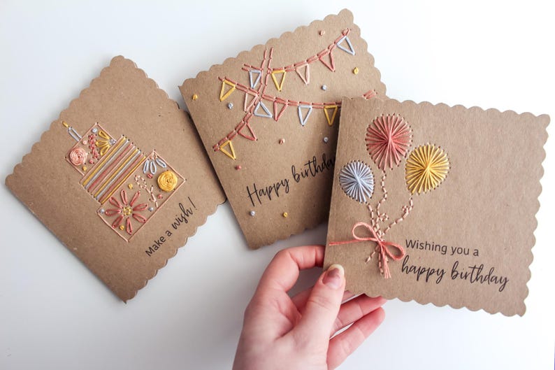 May include: Three handmade birthday cards with scalloped edges. The cards are made of brown cardstock and feature colorful embroidered designs of a cake, bunting, and balloons. Text on the cards reads "Make a wish!" and "Wishing you a happy birthday."