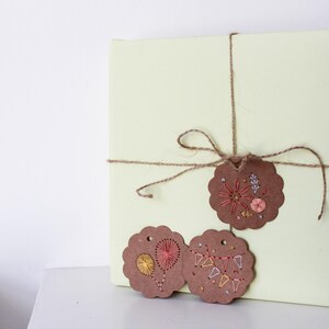 May include: A wrapped present in pale yellow paper, tied with twine and decorated with three brown, scalloped-edge tags. The tags have colourful embroidered designs, including floral and geometric patterns, giving a handcrafted feel to the gift.