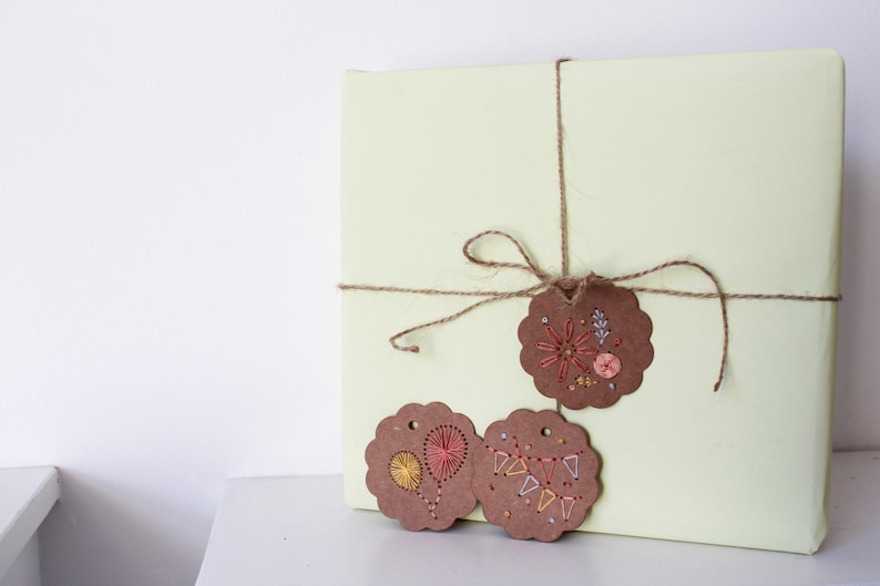 May include: A wrapped present in pale yellow paper, tied with twine and decorated with three brown, scalloped-edge tags. The tags have colourful embroidered designs, including floral and geometric patterns, giving a handcrafted feel to the gift.
