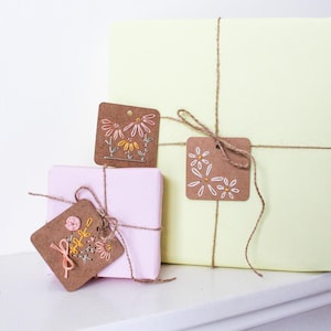 May include: Three wrapped gifts in pastel colors, tied with twine and adorned with embroidered tags. The tags feature floral designs in shades of pink, yellow, and white. The largest gift is pale yellow, while the others are pink.