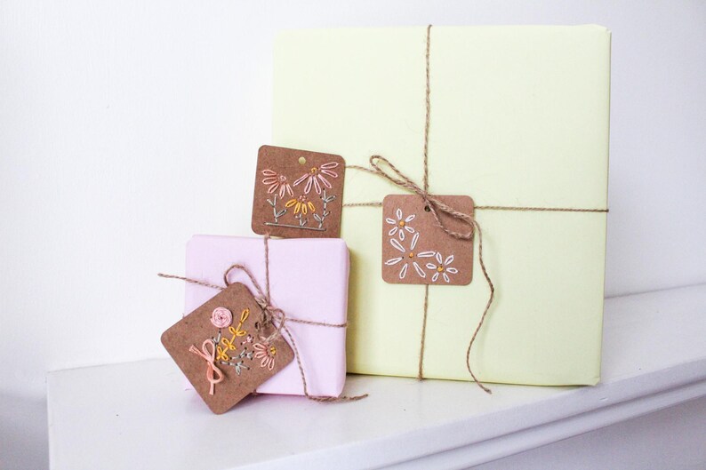 May include: Three wrapped gifts in pastel colors, tied with twine and adorned with embroidered tags. The tags feature floral designs in shades of pink, yellow, and white. The largest gift is pale yellow, while the others are pink.