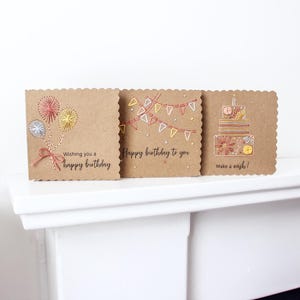 May include: Three birthday cards with scalloped edges are displayed. The cards are made of brown cardstock and feature embroidered designs. One card has fireworks, another has a banner, and the last has a cake. Each card has a birthday message.