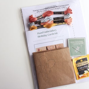 May include: A hand embroidery kit for birthday cards. The kit includes a brown paper card with a pre-punched design of balloons, embroidery floss in yellow, gray, and pink, a needle, and a tea bag.