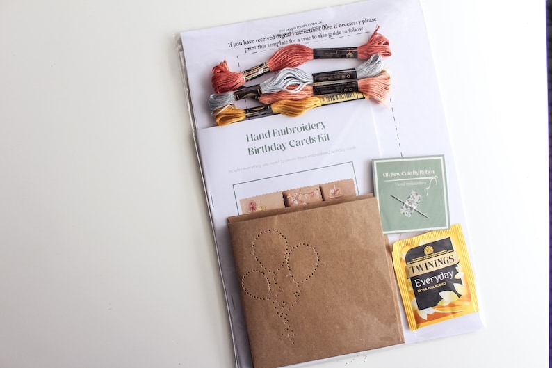 May include: A hand embroidery kit for birthday cards. The kit includes a brown paper card with a pre-punched design of balloons, embroidery floss in yellow, gray, and pink, a needle, and a tea bag.