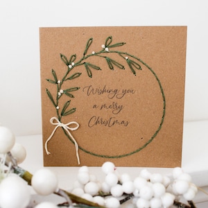 May include: A square, brown Christmas card with a stitched green wreath design and the words "Wishing you a merry Christmas." The wreath is adorned with white beads and a white bow. A sprig of white berries is in the foreground.