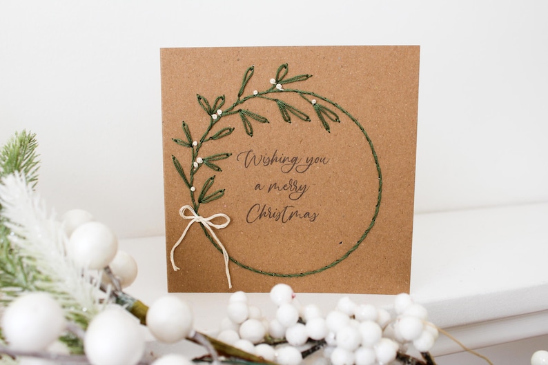 May include: A square, brown Christmas card with a stitched green wreath design and the words "Wishing you a merry Christmas." The wreath is adorned with white beads and a white bow. A sprig of white berries is in the foreground.
