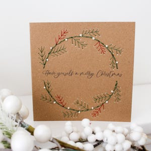 May include: A square, brown Christmas card with a stitched wreath design. The wreath is made of green and red embroidered branches with small white dots. The text "Have yourself a merry Christmas" is written in cursive below the wreath.