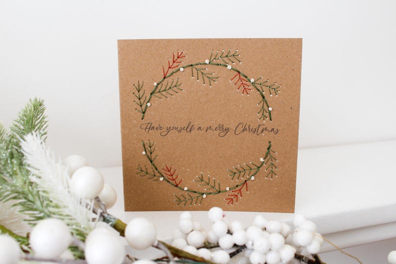 May include: A square, brown Christmas card with a stitched wreath design. The wreath is made of green and red embroidered branches with small white dots. The text "Have yourself a merry Christmas" is written in cursive below the wreath.