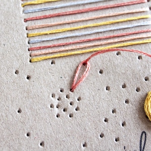 May include: Close-up of a craft project featuring embroidery on a tan surface. The design includes a stitched rectangle with colorful horizontal lines and a yellow circular element. A needle and thread are visible.
