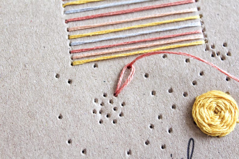 May include: Close-up of a craft project featuring embroidery on a tan surface. The design includes a stitched rectangle with colorful horizontal lines and a yellow circular element. A needle and thread are visible.