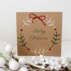 May include: A square, kraft paper Christmas card with a hand-stitched wreath design. The wreath is made of white thread with green leaves and red berries. The words "Merry Christmas" are written in elegant script.