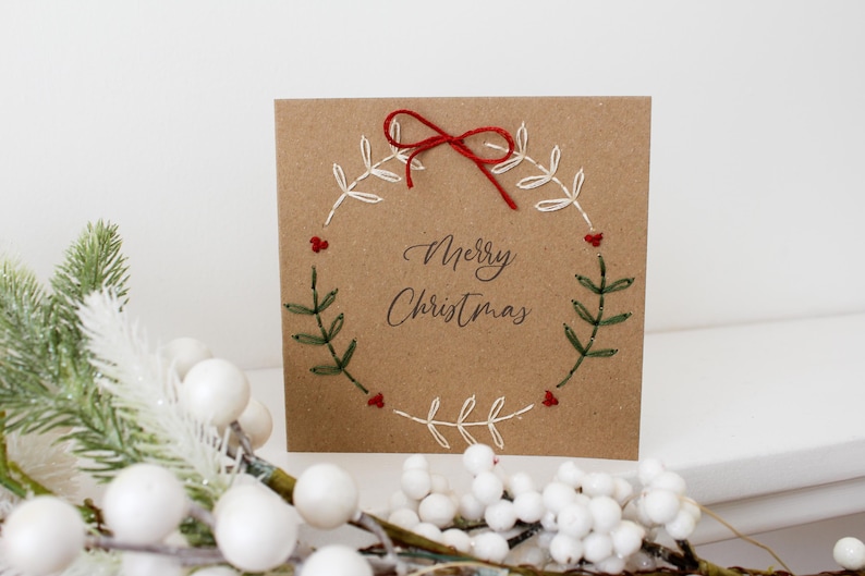 May include: A square, kraft paper Christmas card with a hand-stitched wreath design. The wreath is made of white thread with green leaves and red berries. The words "Merry Christmas" are written in elegant script.