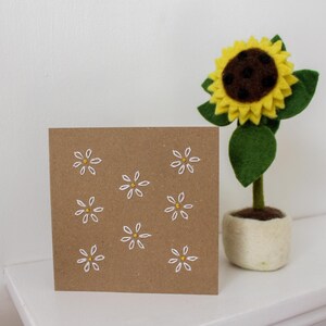 May include: A square, brown card with white and yellow daisy embroidery. A felt sunflower with a yellow bloom, brown center, and green leaves sits in a small, off-white pot. The items are on a white surface.