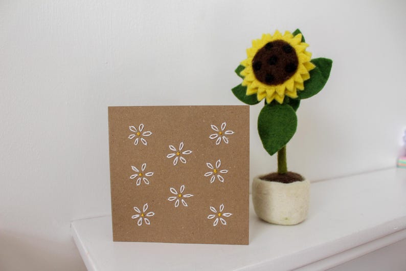 May include: A square, brown card with white and yellow daisy embroidery. A felt sunflower with a yellow bloom, brown center, and green leaves sits in a small, off-white pot. The items are on a white surface.