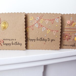 May include: Three handmade birthday cards with a scalloped edge. The cards are made of brown cardstock and feature embroidered designs of balloons, bunting, and a cake. Text on the cards includes "Wishing you a happy birthday", "Happy birthday to you", and "Make a wish!".