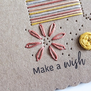 May include: Hand-stitched greeting card with a flower and sun design. The card features a stitched flower in shades of pink and a yellow sun. The phrase "Make a wish" is printed in black.