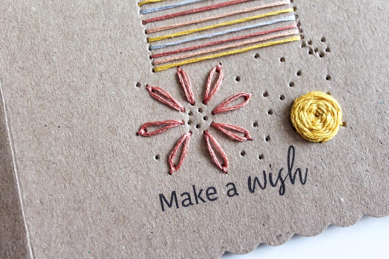 May include: Hand-stitched greeting card with a flower and sun design. The card features a stitched flower in shades of pink and a yellow sun. The phrase "Make a wish" is printed in black.