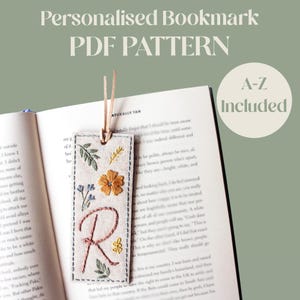 May include: A rectangular fabric bookmark featuring hand-embroidered floral motifs and a large letter R in pink thread. The bookmark has a light tan base, a leather cord at the top, and is placed on an open book. Text reads: Personalised Bookmark PDF PATTERN and A-Z Included.