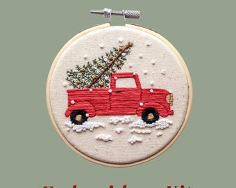 May include: An embroidery kit featuring a red truck carrying a Christmas tree, stitched on a cream-colored fabric. The design includes white snow and is set in a wooden hoop. The text "Embroidery Kit" is displayed in red at the bottom.