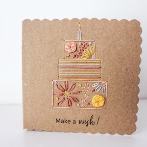 May include: A brown card with a scalloped edge features an embroidered birthday cake with a candle on top. The cake is decorated with flowers and stripes in shades of yellow, orange, and red. The card reads "Make a wish!"