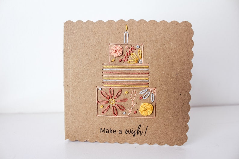 May include: A brown card with a scalloped edge features an embroidered birthday cake with a candle on top. The cake is decorated with flowers and stripes in shades of yellow, orange, and red. The card reads "Make a wish!"