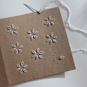 May include: A handmade greeting card with a brown paper cover. The card features white embroidered flower designs. A needle is inserted into the card, with white thread attached, suggesting the card is in progress.