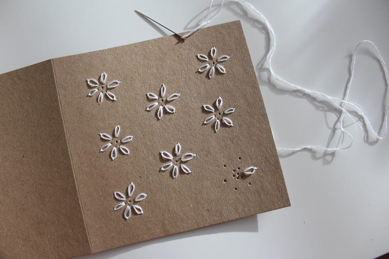 May include: A handmade greeting card with a brown paper cover. The card features white embroidered flower designs. A needle is inserted into the card, with white thread attached, suggesting the card is in progress.