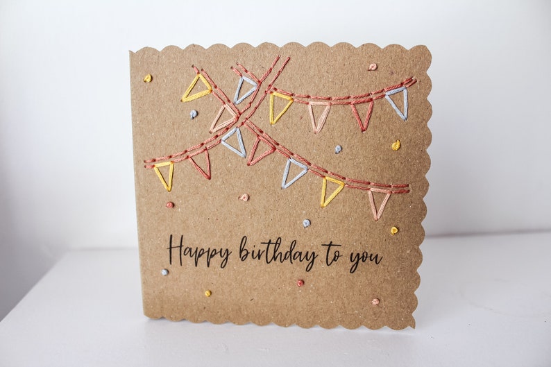 May include: A brown card with a scalloped edge and a hand-stitched design of colorful bunting flags. The card says "Happy birthday to you" in black lettering.