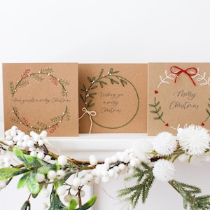 May include: Three square Christmas cards with embroidered wreath designs and festive greetings. The cards are made of brown cardstock. One card says "Have yourself a merry Christmas". Another says "Wishing you a merry Christmas". The third says "Merry Christmas".