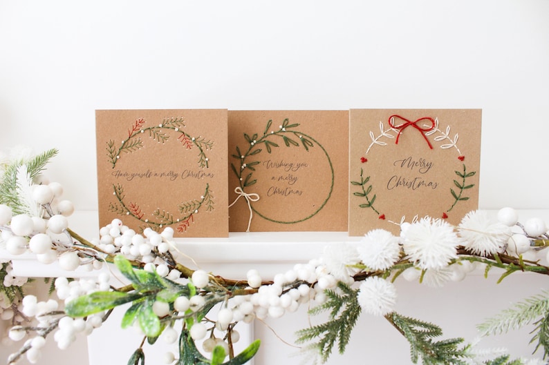 May include: Three square Christmas cards with embroidered wreath designs and festive greetings. The cards are made of brown cardstock. One card says "Have yourself a merry Christmas". Another says "Wishing you a merry Christmas". The third says "Merry Christmas".