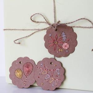 May include: Three brown paper gift tags with embroidered designs. One tag features a flower, another a heart-shaped design, and the third a banner. The tags are tied with twine to a light green box, creating a rustic, handmade aesthetic.