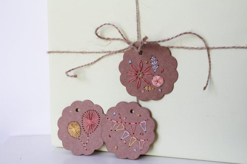 May include: Three brown paper gift tags with embroidered designs. One tag features a flower, another a heart-shaped design, and the third a banner. The tags are tied with twine to a light green box, creating a rustic, handmade aesthetic.