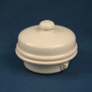 May include: A cream-colored ceramic butter dish with a lid and a small knob on top. The dish has a round base and a decorative design on the side. The background is a solid dark blue.